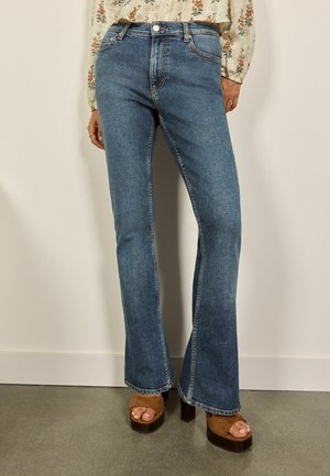 Kvinde iført blå højtaljede bootcut-jeans og brune platformsandaler, stående mod en ensfarvet lys væg og gulv.