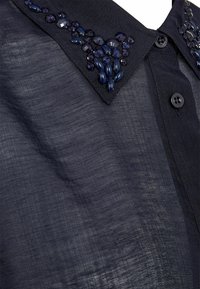 Tume sinine läbipaistev pluus, millel on nööbid ees ja krae, mida kaunistavad klaaspärlid tumedas sinises värvis klastrite mustrites.