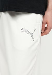 Κοντινό πλάνο λευκού παντελονιού με λογότυπο Puma από στρας στο μηρό, φορεμένο με μαύρο πουκάμισο και ένα χαλαρό χέρι στο πλάι.