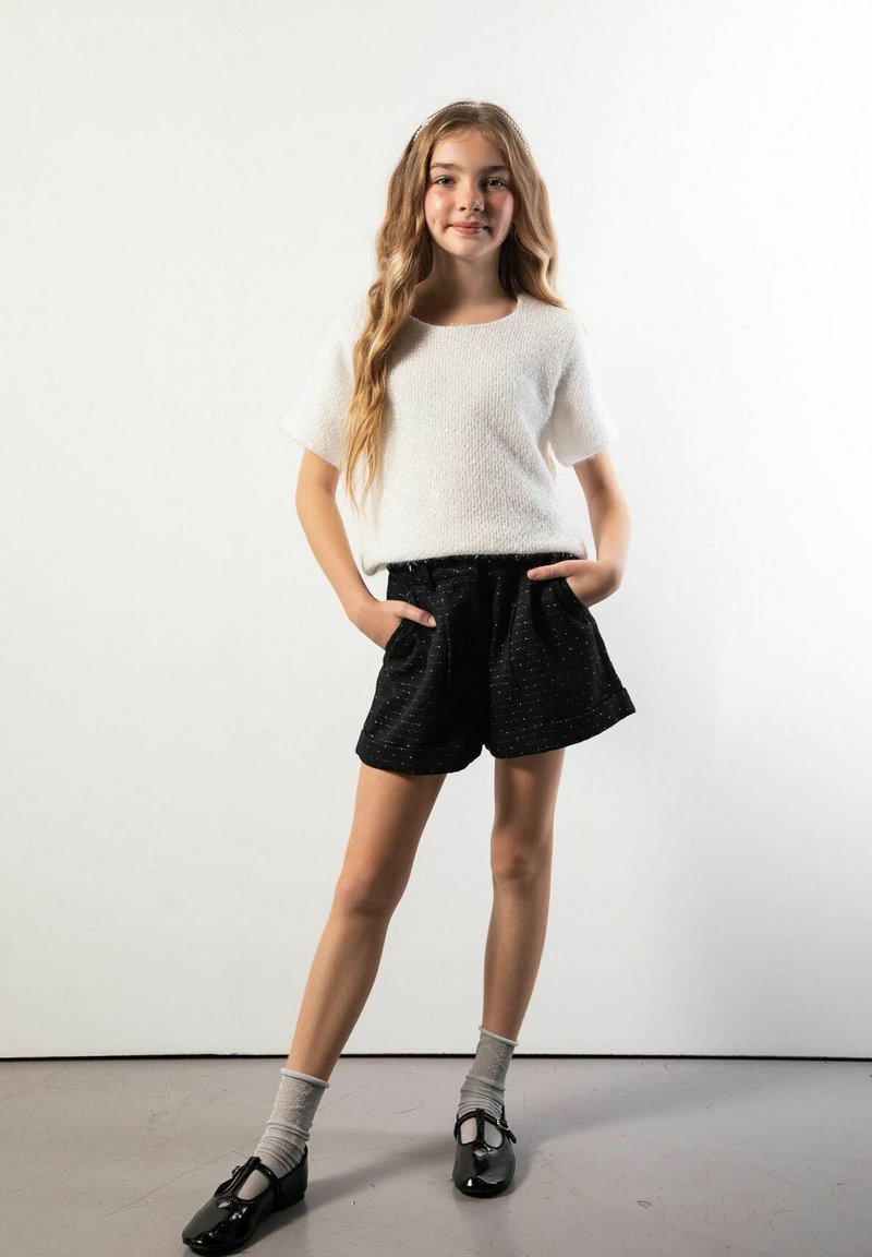 Biały top z krótkim rękawem z teksturowanej tkaniny, zestawiony z czarnymi szortami o subtelnym wzorze w kropki, szarymi skarpetami i czarnymi butami.