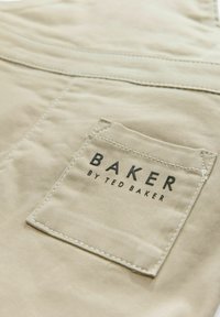 Beige kangasta, jossa on näkyvä tasku. Taskussa on mustalla tekstillä kirjoitettu "BAKER" ja sen alapuolella pienemmällä fontilla "by TED BAKER", valkoisella ommeltuna.