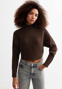 Maglione marrone a costine con collo alto e design cropped, abbinato a jeans blu sbiaditi a vita alta. Texture morbida e vestibilità rilassata.