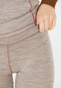 Lichtbruine, gestructureerde leggings met een gladde oppervlakte, voorzien van contraststiksel en een comfortabele tailleband. Hand die de stof vasthoudt.
