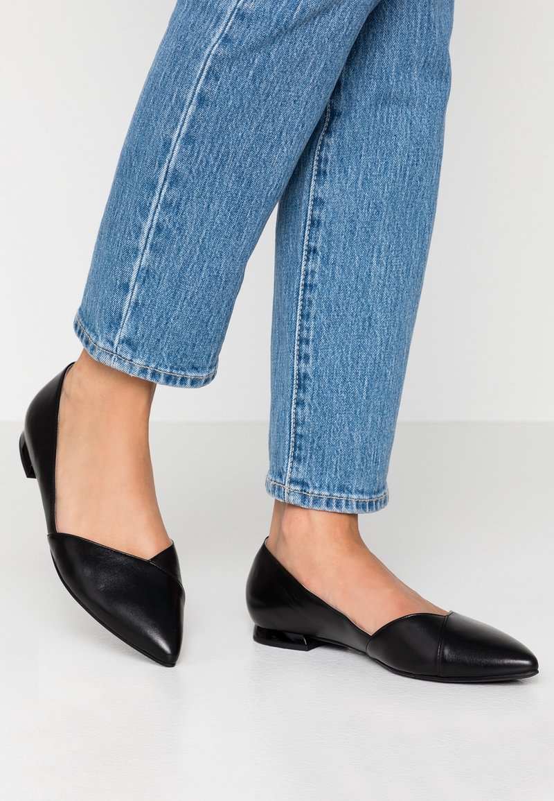 Sorte læderballerinaer med spids tå og v-formet design, der har en lav hæl og glat overflade, båret med blå denim jeans.