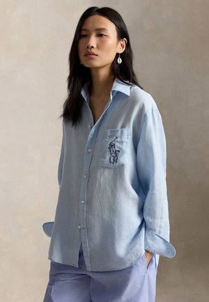 Frau mit langen dunklen Haaren trägt ein hellblaues Hemd mit Knöpfen, auf dessen Tasche ein gestickter Polospieler abgebildet ist, und passende hellblaue Hosen.