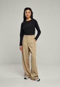 Chemise noire à manches longues avec un col rond, associée à un pantalon large beige et des chaussures de sport grises. Design simple, texture du tissu visible.