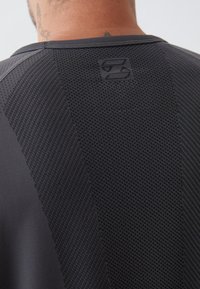 Herren Sportshirt, schwarz mit strukturierten Mesh-Einsätzen. Mit Rundhalsausschnitt und einem Logo auf der Rückseite. Glattes und atmungsaktives Material.