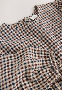 Blúzka s károvaným vzorom v béžovej, bordovej a námorníckej modrej; má okrúhly výstrih a nariasené detaily, vyrobená z mäkkého materiálu.
