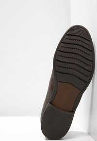 Semelle d'une chaussure habillée marron foncé avec des motifs de grip texturés, inclinée contre un fond blanc aux angles nets.