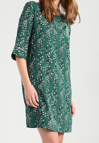 Vestido verde hecho de una tela suave, con un patrón floral en blanco, amarillo y púrpura. Tiene un corte holgado y mangas de tres cuartos.