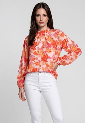 Frau mit langen dunklen Haaren, die eine pinke Bluse mit orangefarbenem Blumenmuster und weiße Hose trägt, steht mit einer Hand auf der Hüfte.