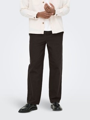 Mann trägt schwarze gerade geschnittene Hose, schwarze Lederloafer, weißes Hemd und beige Jacke mit Knöpfen, steht mit verschränkten Händen.