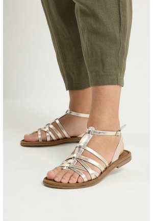Voeten met zilverkleurige, platte sandalen met bandjes, gecombineerd met olijfgroene, cropped broeken tegen een effen witte achtergrond.