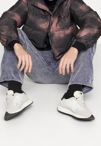 Persona sentada con las piernas abiertas, vistiendo una chaqueta de tie-dye oscura, pantalones de pana azul, calcetines negros y zapatillas blancas de New Balance.