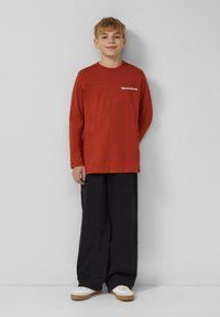 Chłopiec stoi, ubrany w rdzawą koszulę z długim rękawem z napisem, szerokie czarne spodnie i białe sneakersy, na tle jednolitej szarej ściany.