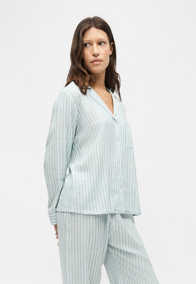 Femme aux longs cheveux bruns portant un haut de pyjama rayé bleu clair et blanc à manches longues avec boutons et un pantalon assorti, debout sur fond blanc.