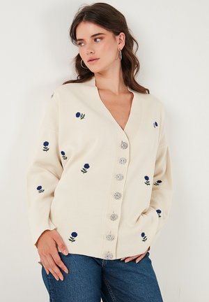 Cardigan crema con scollatura a V, caratterizzato da ricami floreali blu e sei bottoni argentati. Tessuto morbido e vestibilità rilassata.