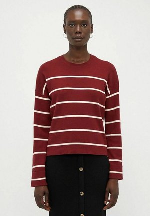 Donna che indossa un maglione rosso con strisce orizzontali bianche e una gonna nera con bottoni, in piedi contro uno sfondo chiaro e uniforme.