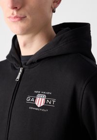 Crna zip-up hoodica od mekanog materijala s grafičkim dizajnom koji sadrži tekst "New Haven" i "GAIANT", uz grb s crvenim, bijelim i plavim dezenima.