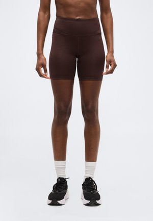 Άτομο που φοράει σκούρα καφέ εφαρμοστά σορτς ποδηλασίας, λευκές κάλτσες και μαύρα αθλητικά παπούτσια, στήνεται μπροστά σε απλό ανοιχτό φόντο.