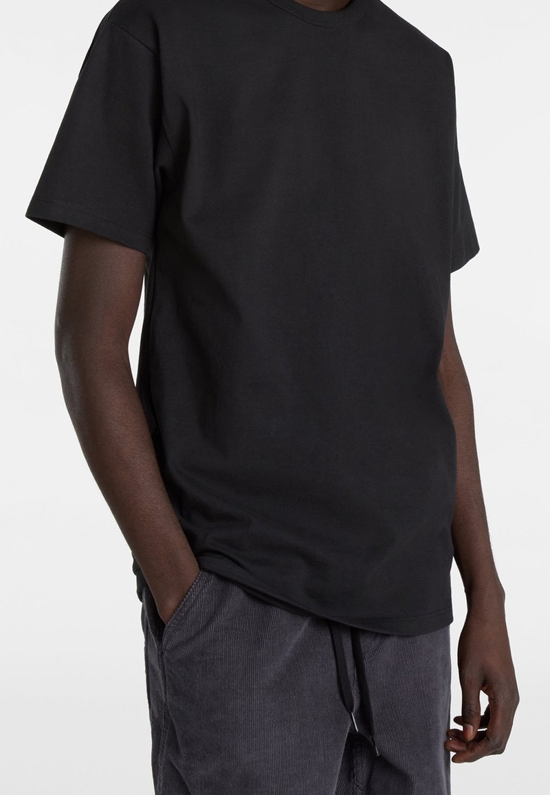 Personne portant un t-shirt noir uni à manches courtes avec une main dans la poche d’un pantalon en velours côtelé gris foncé.