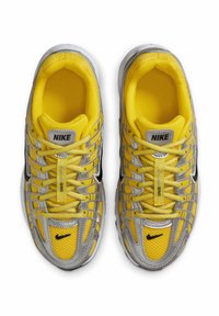 Nike-Sneaker mit einem oberen Teil aus gelbem Netzgewebe, silbernen Akzenten, schwarzem Logo und strukturiertem Profil. Schnürsenkel und Markendetails auf der Zunge vorhanden.