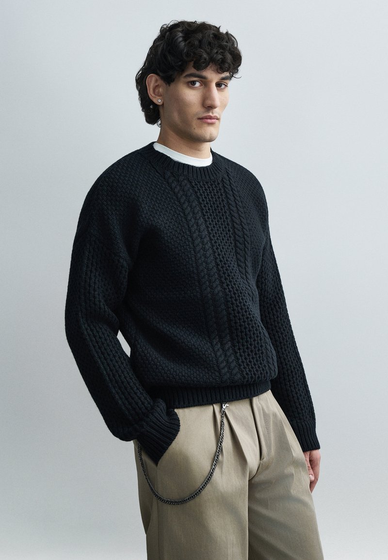 Άνδρας με κοντά σγουρά μαλλιά, φοράει μαύρο πουλόβερ με υφή και μπεζ παντελόνι με αλυσίδα, στέκεται με το ένα χέρι στην τσέπη σε απλό φόντο.