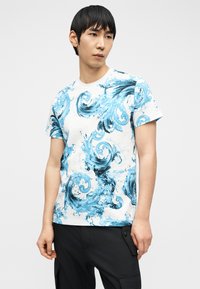 Άνδρας με μαύρα μαλλιά που φοράει άσπρο T-shirt με μπλε σχέδια πιτσιλιών νερού και μαύρο παντελόνι, στέκεται μπροστά σε απλό ανοιχτό φόντο.