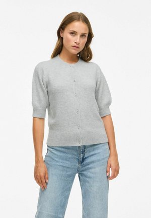 Cardigan grigio a maniche corte realizzato in morbido tessuto a maglia, con un collo rotondo e chiusura frontale con cinque bottoni. Abbinato a jeans blu chiaro.