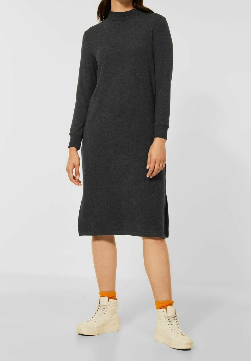 Langärmliges, dunkelgraues Strickkleid mit hohem Ausschnitt und seitlichen Schlitzen. Kombiniert mit cremefarbenen High-Top-Sneakern und orangen Knöchelsocken.