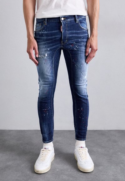 Schmal geschnittene dunkelblaue Jeans mit Farbspritzern und Abnutzungen, getragen mit weißen Sneakers und einem weißen Hemd, stehend auf einem grauen Boden.