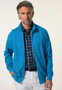 Giacca a zip azzurro chiaro con colletto alto, realizzata in tessuto lisci; indossata sopra una camicia a quadri blu e verde e pantaloni bianchi.