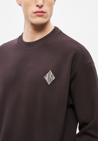 Καφέ φούτερ με ριμπ ύφασμα στο λαιμό, υλικό με υφή και κεντημένο λευκό λογότυπο σε σχήμα ρόμβου στο στήθος. Κοντινή άποψη.