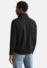 Zwarte fleece trui met hoge kraag, geribbelde manchetten en tailleband. Draagt een lichtblauwe denim jeans, gezien vanaf de achterkant.