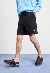 Black athletic shorts with a drawstring waist and a white Jordan logo, paired with black leather clogs featuring a strap and yellow stitching.