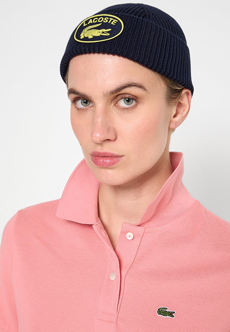 Hellrosa Poloshirt mit strukturiertem Finish und einem Kragen mit zwei Knöpfen. Navy-Rippmütze mit einem gelben Lacoste-Logo-Patch.