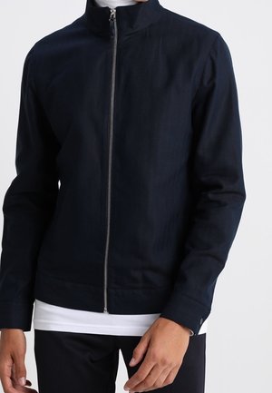 Mężczyzna w granatowej kurtce zapinanej na zamek na białą koszulę i czarne spodnie, stojący na tle jasnego, jednolitego tła.