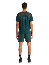 Mand stående set forfra iført en mørkegrøn og grå kortærmet skjorte og matchende shorts, sorte sokker og sorte sneakers.