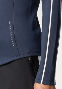 Námořnický modrý sportovní top s dlouhým rukávem a bílými bočními pruhy, vyrobený z hladkého, elastického materiálu. Obsahuje boční zip a detail s logem.