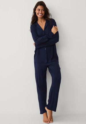 Kvinde smilende, stående barfodet, iført navyblå langærmet knap-op bluse og matchende løse bukser mod en ensfarvet lys baggrund.