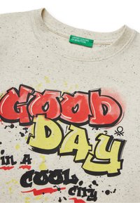 Helles beige Baumwoll-T-Shirt mit gesprenkelter Textur, das einen auffälligen grafischen Druck in Rot, Gelb und Schwarz mit der Aufschrift "GUTER TAG in einer coolen Stadt" aufweist.