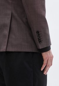 Maßgeschneiderte Jacke in Helllila mit einer strukturierten Webart, ausgestattet mit sechs schwarzen Knöpfen am Ärmelbündchen. Kombiniert mit schwarzen Hosen.