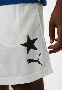 Hvite sports shorts med en glatt tekstur, som har en svart stjernegrafikk og en svart Puma-logo. Blå midjevisning øverst.