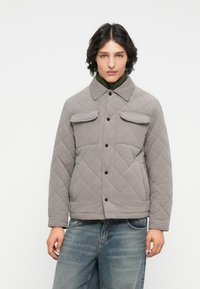 Veste grise matelassée avec un col, comportant deux poches poitrine et des boutons noirs, portée par-dessus une couche intérieure verte. Un jean en denim est visible en dessous.