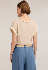 Blusa beige a maniche corte con risvolti, dal tessuto liscio e con scollatura a barchetta. Abbinata a pantaloni azzurri a vita alta con cintura beige.