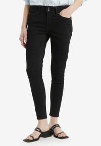 Jeans negros ajustados con una cintura de media altura, bolsillos delanteros y una textura de denim suave, combinados con sandalias negras de tacón cuadrado.