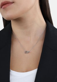 Collana d'argento con un design a cuori intrecciati e a maglie su una sottile catena, indossata sopra una maglietta bianca e un blazer nero.