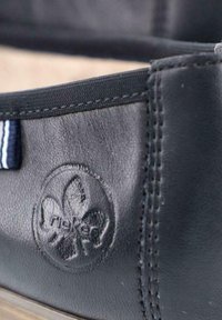 Schwarze Leder-Hausschuhe mit rundem Logo, kontrastierenden Nähten und einem kleinen gestreiften Stoffdetail entlang des Randes.
