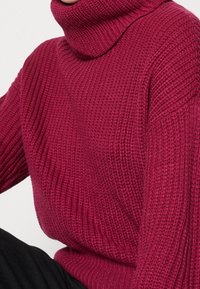 Burgundy neulepaita, jossa on teksturoitu ribbikuvio ja väljä istuvuus, pitkät hihat ja paksu kaula-aukko.