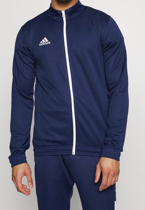Homme portant une veste de survêtement Adidas bleu marine avec une fermeture éclair blanche et un logo sur la poitrine gauche, debout devant un fond uni.